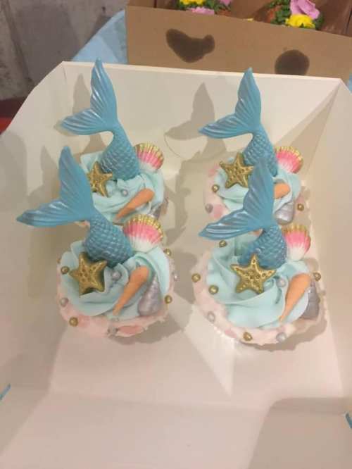 Four mermaid-themed cupcakes with blue tails, seashells, and starfish decorations on a light blue frosting base.