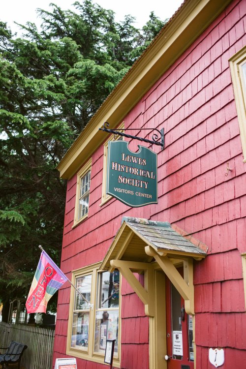Sign for the Lewes Historical Society Visitors Center on a red shingled building, with a flag nearby.