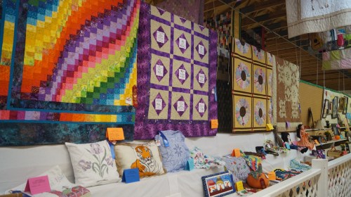Colorful quilts and decorative pillows displayed on a wall, with a vendor sitting behind a table in a craft fair setting.