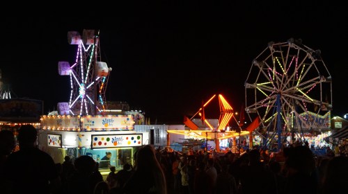 A vibrant night scene at a carnival with illuminated rides and food stalls, bustling with people.