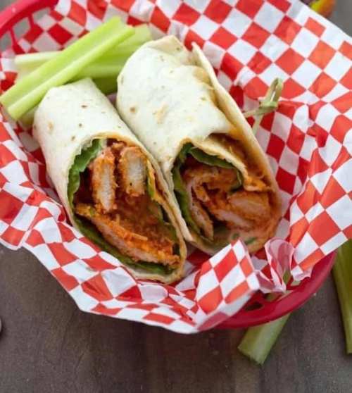 Two chicken wraps with lettuce and sauce in a red and white checkered basket, accompanied by celery sticks.