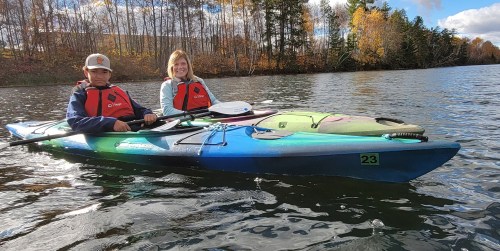A boy and a woman in life jackets sit in a colorful kayak on a calm lake surrounded by autumn trees.