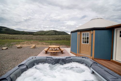 A hot tub beside a yurt, with a picnic table and scenic mountains in the background under a cloudy sky.