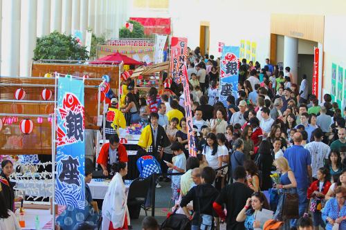 A bustling festival scene with colorful banners, food stalls, and a large crowd enjoying the festivities indoors.