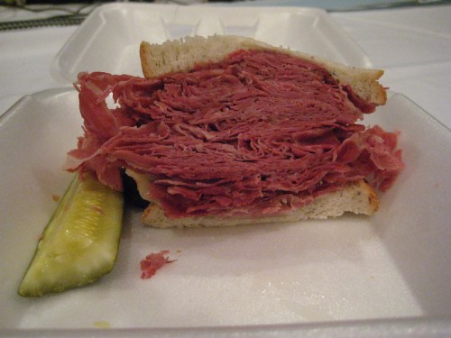 A close-up of a stacked corned beef sandwich on white bread, accompanied by a pickle slice on a foam takeout container.