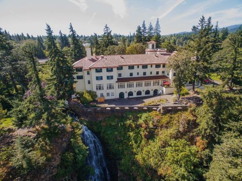 Aerial view of a large, historic building surrounded by trees and a waterfall, set against a clear blue sky.