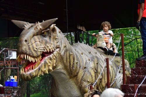A child sits on a large dinosaur statue, smiling, with a forest backdrop in a playful setting.