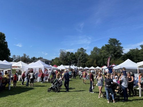 A bustling outdoor market with white tents, vendors, and visitors enjoying a sunny day in a green park.