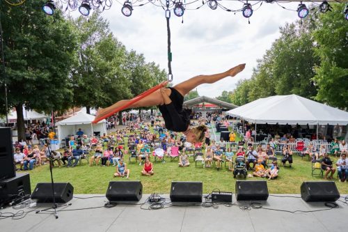 Aerial performer in mid-air above a crowd at an outdoor festival, with tents and spectators in the background.