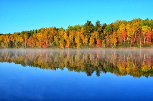 A serene lake reflecting vibrant autumn trees under a clear blue sky, with mist rising from the water's surface.
