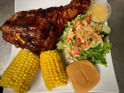 A plate featuring a grilled turkey leg, corn on the cob, a fresh salad, and a dinner roll with a side of sauce.
