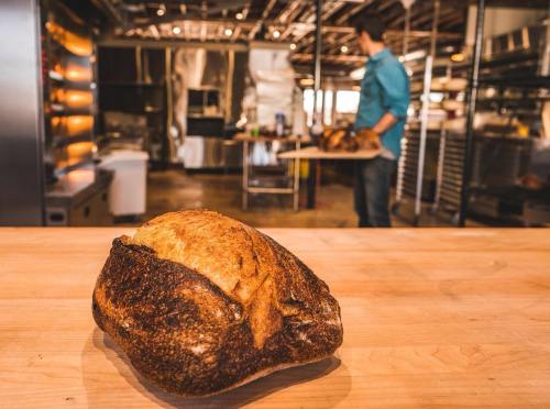 A freshly baked loaf of bread on a wooden table, with a baker in the background preparing more bread.