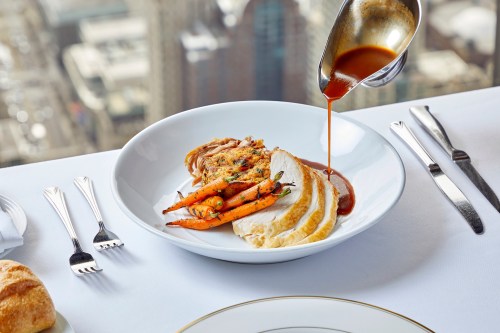 A plate of sliced chicken with carrots and sauce, set on a table with a city view in the background.