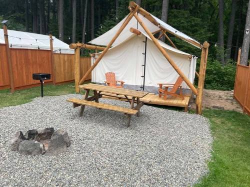A cozy outdoor campsite with a tent, picnic table, and fire pit surrounded by trees and wooden fencing.