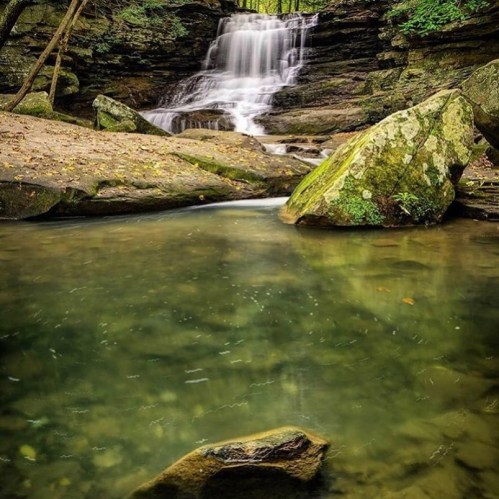 A serene waterfall cascades over rocks into a calm pool, surrounded by lush greenery and natural stone formations.