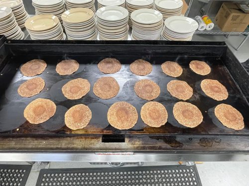 A griddle with multiple pancakes cooking, surrounded by stacks of plates in a kitchen setting.