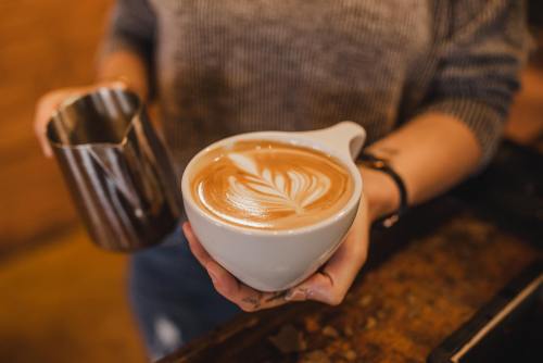 A person holds a large cup of latte art, with a milk pitcher in the other hand, in a cozy café setting.