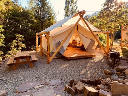 A cozy glamping tent with wooden supports, surrounded by trees and a picnic table on a gravel area.