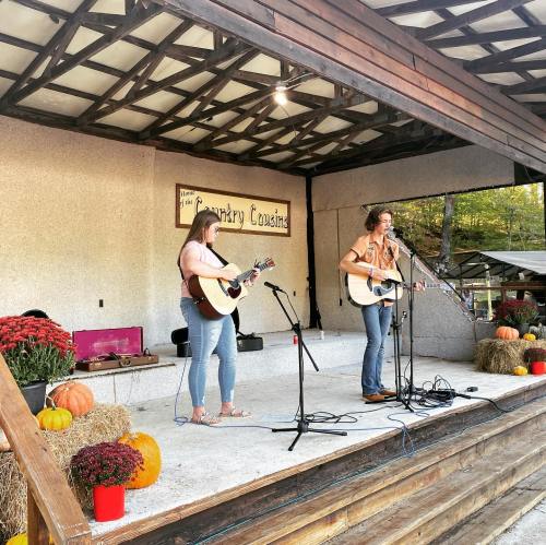 Two women perform on stage with guitars, surrounded by pumpkins and hay bales, in a cozy outdoor setting.
