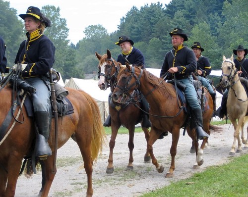A group of mounted riders in historical military uniforms, riding along a path near tents and trees.