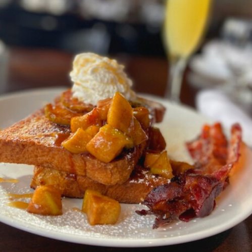 A plate of French toast topped with caramelized apples, whipped cream, and crispy bacon, with a mimosa in the background.