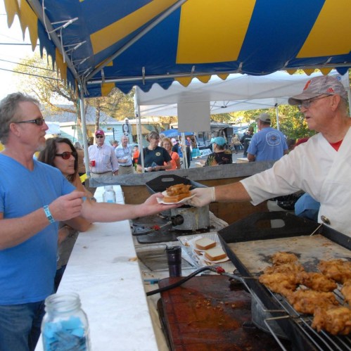 A vendor hands a plate of fried food to a customer at a lively outdoor festival with tents and people in the background.