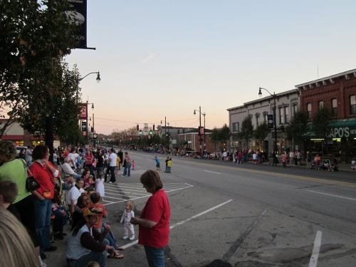 A crowd gathers along a street lined with shops, waiting for a parade at sunset.