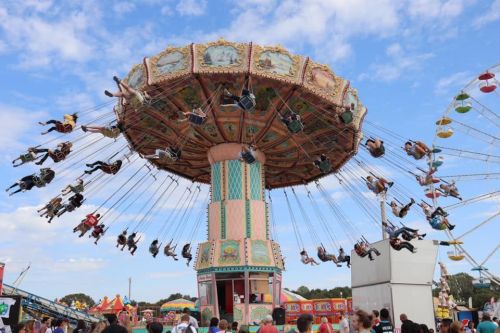 A colorful swing ride at a fair, with people enjoying the view as they soar above a crowd under a blue sky.