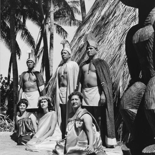 A group of six individuals in traditional attire poses outdoors, surrounded by palm trees and a thatched structure.