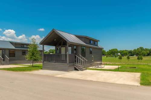 A modern gray cabin with a metal roof, porch, and surrounding green grass under a clear blue sky.