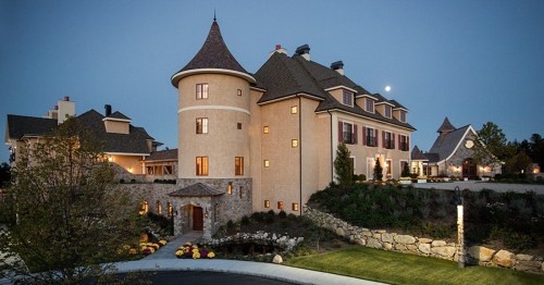 A large, elegant castle-like home with turrets, surrounded by landscaped gardens, illuminated at dusk.