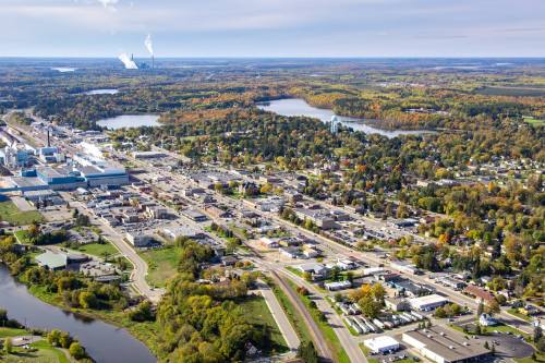Aerial view of a small town surrounded by greenery and water, with industrial buildings and autumn foliage in the background.