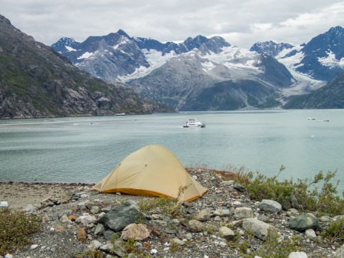 A yellow tent on a rocky shore, with mountains and a glacier in the background and a boat on the calm water.