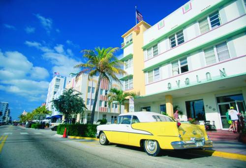 A vintage yellow car parked on a sunny street lined with palm trees and colorful Art Deco buildings.