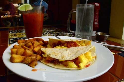 A plate with two breakfast tacos, seasoned potatoes, and a drink on a colorful tiled table.