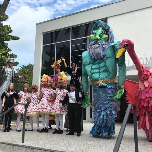A colorful group in costumes, including a giant blue figure, a flamingo, and performers in polka-dot dresses, poses outside a building.