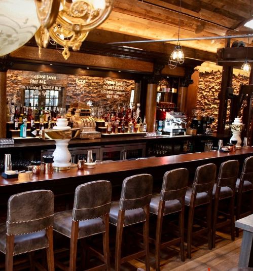 A cozy bar interior featuring a wooden counter, high stools, and shelves stocked with various bottles. Warm lighting enhances the ambiance.