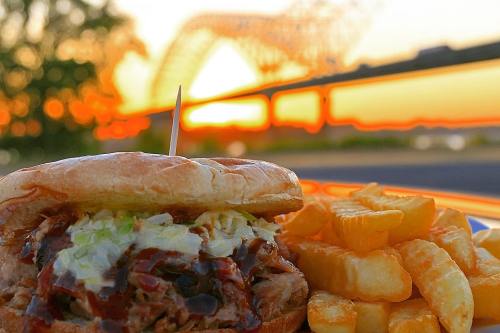 A close-up of a pulled pork sandwich with lettuce and barbecue sauce, served with crinkle-cut fries and a sunset backdrop.