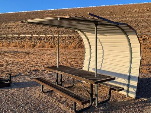 A metal picnic shelter with a table and benches, set against a dry, rocky landscape under a clear blue sky.