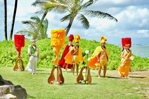 A group of performers in colorful costumes stands on a grassy area by the beach, with palm trees and ocean in the background.
