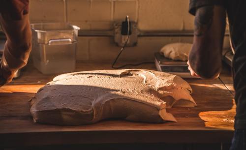 Two hands work with a large, smooth dough on a wooden surface, with tools and containers in the background.