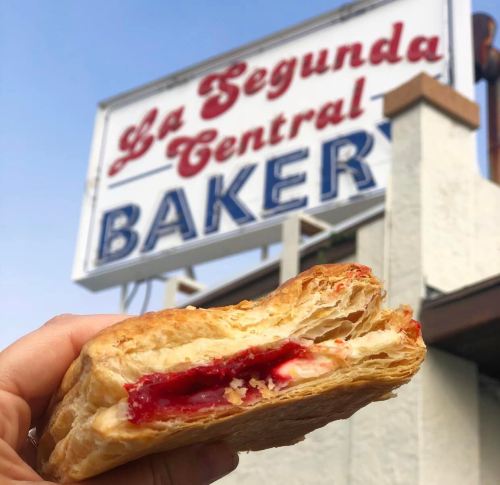 A hand holds a flaky pastry filled with red fruit filling, with a bakery sign in the background.
