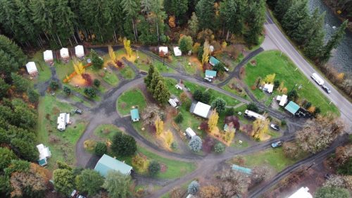 Aerial view of a campground with cabins, RVs, and autumn trees along winding paths near a road and river.