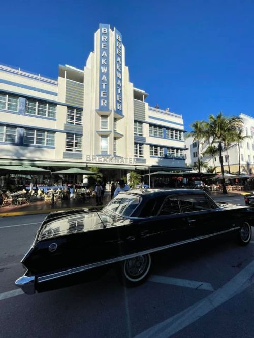 A classic black car parked in front of the Breakwater Hotel, with palm trees and a clear blue sky in the background.