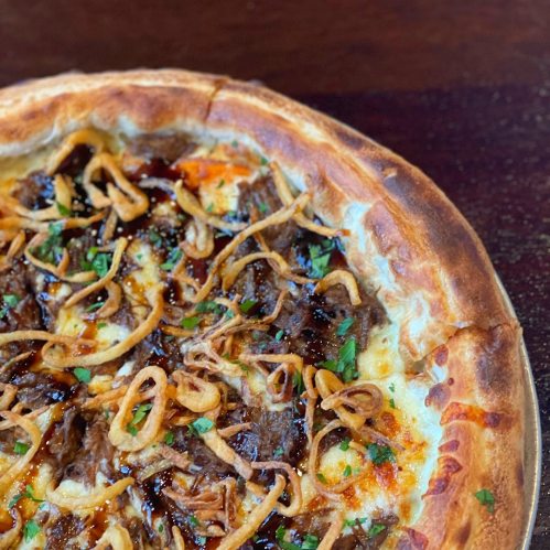 A close-up of a pizza topped with shredded beef, crispy onions, and herbs on a golden crust.