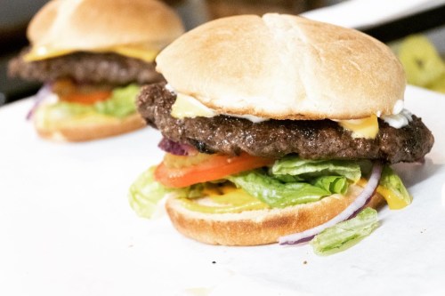 Two juicy burgers with cheese, lettuce, tomato, onion, and mayonnaise on soft buns, served on a white surface.