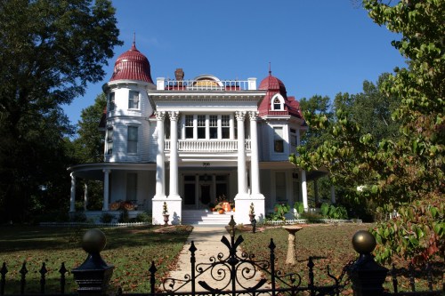 A grand Victorian-style house with red turrets, white siding, and a lush garden, framed by a decorative black fence.