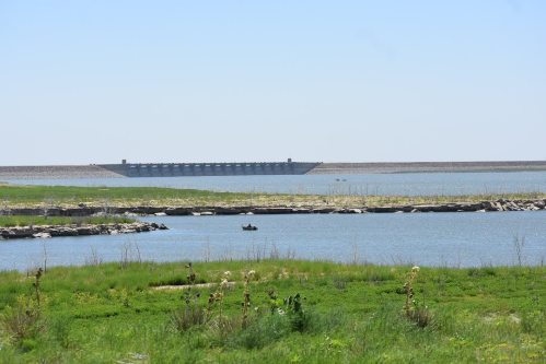 A serene landscape featuring a calm lake, grassy shorelines, and a distant dam under a clear blue sky.