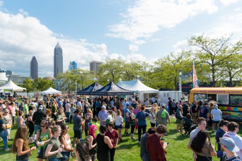 A lively outdoor festival with crowds, food tents, and city skyline in the background under a partly cloudy sky.