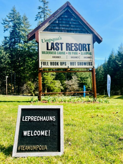 Sign for Umpqua's Last Resort with "Leprechauns Welcome!" on a chalkboard in front of a forested area.
